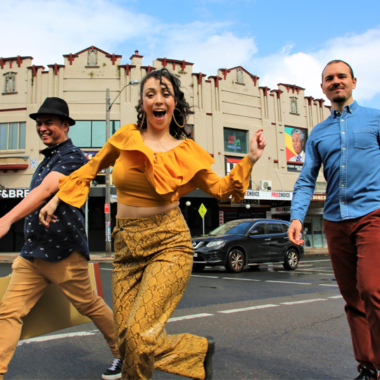 Three people in colourful clothing walk and leap across Marrickville Road with Lazybones music venue in the background